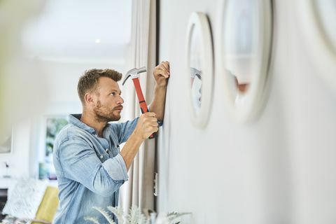 sidebillede af ung mand, der hamrer søm på hvid væg smuk mand holder værktøj i hjemmet, han er hjemme