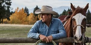 kevin costner i yellowstone ved siden af ​​en hest, der læner sig op af et hegn med reb i hænderne iført en falmet blå denimskjorte og beige cowboyhat