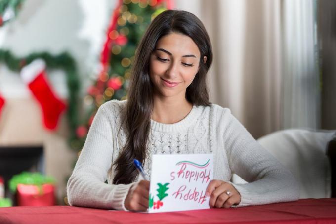 en smilende ung kvinde sidder ved et bord i sin dekorerede stue og kigger koncentreret ned, mens hun skriver et brev til en ven til jul