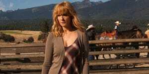 kelly reilly som beth dutton i en brun hvid og rød plaid kjole med brun cardigan stående foran en hesteindhegning