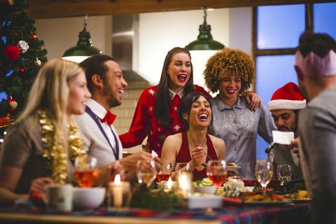 en gruppe venner griner, mens de sidder rundt om et bord og spiser og drikker i julen, en mand læser en vittighed fra en juleknækker festhatte og juletrøjer er brugt